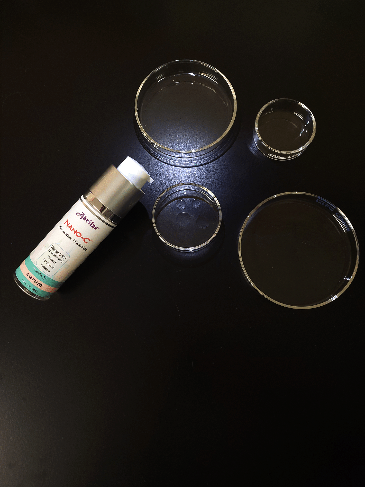 Nano-C vitamin C serum drops onto a glass dish on a black lab counter. There are 4 glass dishes of varying sizes.